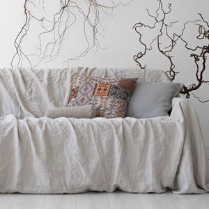 K&ouml;nnte beinhalten: Ein hellfarbenes Leinensofa, das mit einer passenden H&uuml;lle drapiert ist. Dekorative Kissen in Grau-, Braun- und Beiget&ouml;nen liegen auf dem Sofa. Die Wand hinter dem Sofa ist wei&szlig;, mit braunen &Auml;sten als Dekoration.
