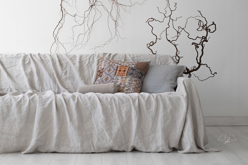 K&ouml;nnte beinhalten: Ein hellfarbenes Leinensofa, das mit einer passenden H&uuml;lle drapiert ist. Dekorative Kissen in Grau-, Braun- und Beiget&ouml;nen liegen auf dem Sofa. Die Wand hinter dem Sofa ist wei&szlig;, mit braunen &Auml;sten als Dekoration.