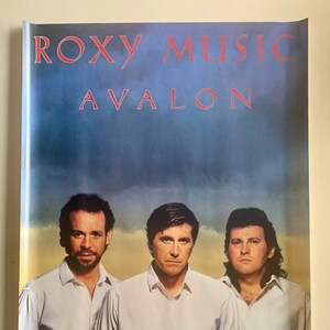 May include: A vintage poster for the band Roxy Music's album Avalon. The poster features a photo of the band members in white shirts and dark pants against a blue and yellow sky. The band's name and album title are printed in red letters at the top of the poster.