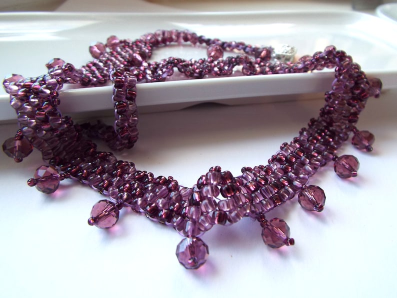 May include: A purple beaded necklace with a fringe of dangling beads. The necklace is made of small, faceted beads that are strung together in a delicate pattern.