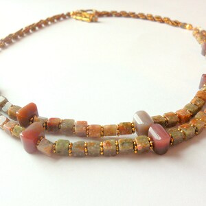 May include: A double-strand necklace made with small, square, multi-colored beads. The beads are a mix of brown, green, and red. The necklace is accented with gold beads.