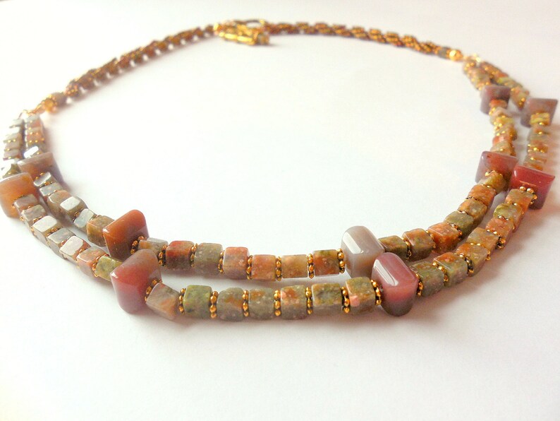 May include: A double-strand necklace made with small, square, multi-colored beads. The beads are a mix of brown, green, and red. The necklace is accented with gold beads.