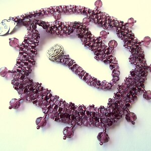 May include: A purple beaded necklace with a silver charm that says "Love is All". The necklace has a fringe of beads that hang down from the bottom.