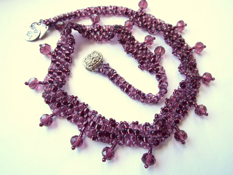 May include: A purple beaded necklace with a silver charm that says "Love is All". The necklace has a fringe of beads that hang down from the bottom.