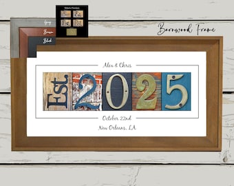 May include: A framed artwork with a brown wooden frame. The artwork features the text "Est 2025" in various colors and textures, along with the names "Alex & Chris", the date "October 22nd", and the city "New Orleans, LA".
