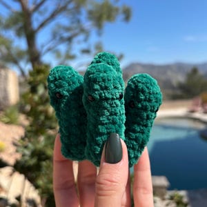May include: Three green plush toys shaped like a pickle with embroidered eyes. The toys are held in a person's hand.
