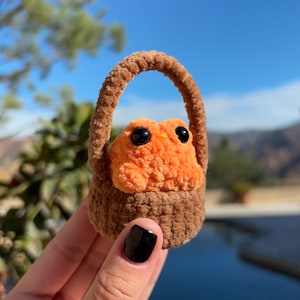 Könnte beinhalten: Ein kleines orangefarbenes Häkeltier mit großen schwarzen Augen sitzt in einem braunen Häkelkorb.