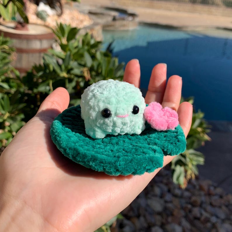 Pu&ograve; includere: Una rana all'uncinetto verde menta fatta a mano con occhi neri e un sorriso rosa, seduta su una foglia di ninfea verde. Un piccolo cuore rosa &egrave; accanto alla rana. La rana e la foglia sono realizzate in morbido filato di peluche.