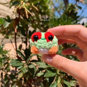 Pu&ograve; includere: Una piccola rana all'uncinetto fatta a mano. La rana ha un corpo verde chiaro, occhi rossi e zampe arancioni. La rana &egrave; tenuta nella mano di una persona su uno sfondo di fogliame verde.