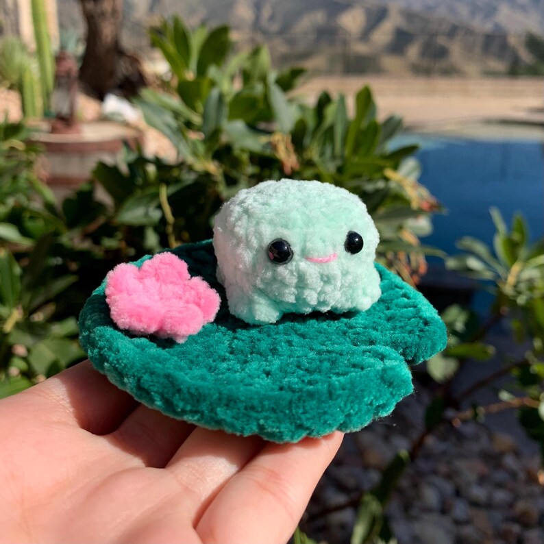 Pu&ograve; includere: Una rana all'uncinetto fatta a mano in verde chiaro, con occhi neri e un sorriso rosa, seduta su una foglia di ninfea verde con un fiore rosa. Il peluche &egrave; tenuto in mano, con uno sfondo sfocato all'aperto.