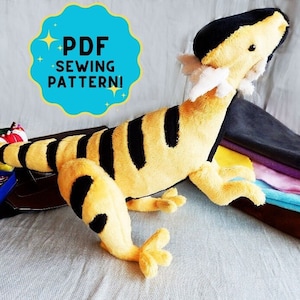 May include: A plush toy lizard in yellow and black stripes, with a black head and tail. The toy has white accents and is lying on a surface. A teal circle with the text "PDF SEWING PATTERN!" is in the upper left.