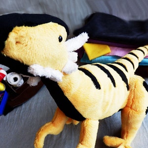 May include: A plush toy dinosaur in yellow and black stripes, with a black head and white accents. The toy is on a gray surface, with sewing supplies and fabric scraps in the background.