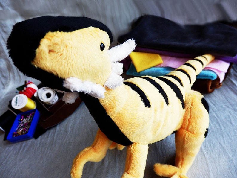 May include: A plush toy dinosaur in yellow and black stripes, with a black head and white accents. The toy is on a gray surface, with sewing supplies and fabric scraps in the background.