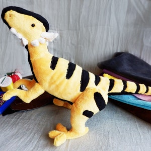 May include: A plush toy dinosaur in a standing pose, featuring a yellow body with black stripes. The dinosaur has a black head and a white frill around its neck. The toy is approximately 12 inches tall.