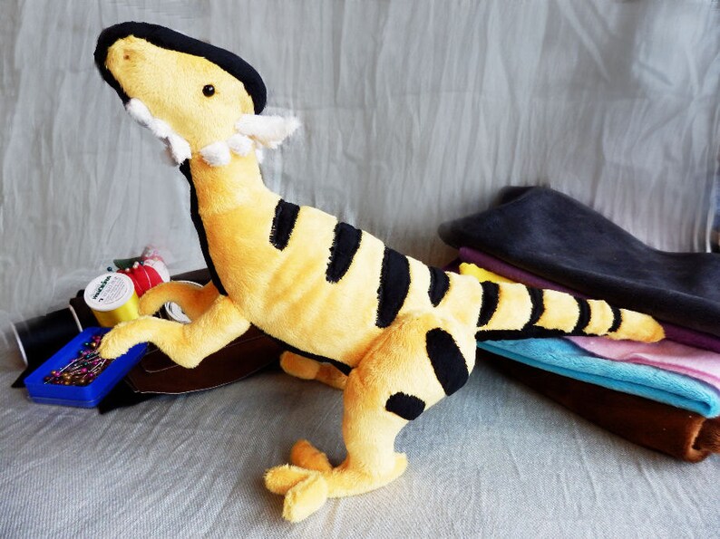 May include: A plush toy dinosaur in a standing pose, featuring a yellow body with black stripes. The dinosaur has a black head and a white frill around its neck. The toy is approximately 12 inches tall.