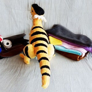 May include: A plush toy tiger with black stripes on a yellow body, a black cap, and a white ruffled collar. Sewing supplies, including thread, pins, and fabric scraps, are arranged nearby. The toy is approximately 12 inches tall.