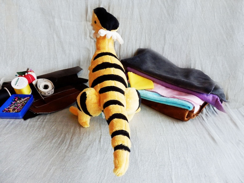 May include: A plush toy tiger with black stripes on a yellow body, a black cap, and a white ruffled collar. Sewing supplies, including thread, pins, and fabric scraps, are arranged nearby. The toy is approximately 12 inches tall.