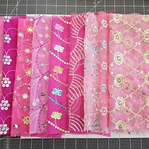 Op de afbeelding: Een verzameling roze stoffen met diverse geborduurde en pailletten patronen. De stoffen hebben bloemen-, vlinder- en geometrische ontwerpen, met gouden, zilveren en blauwe accenten. De stoffen zijn naast elkaar geplaatst.