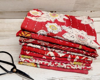 May include: A stack of red kantha fabric squares with white floral patterns. The fabric is folded and stacked, with the top square showing a floral design. The fabric is stitched together with white thread. The fabric is on a white wooden surface. The text "Amour Fabrics" is visible on the top square. A pair of black scissors is in the foreground.