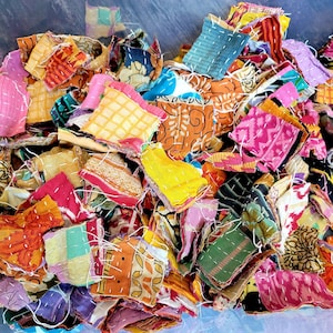 May include: A collection of colorful fabric scraps in a clear plastic container. The scraps are various sizes and shapes, with a variety of patterns and colors, including pink, yellow, orange, and blue. Each piece is stitched with white thread.