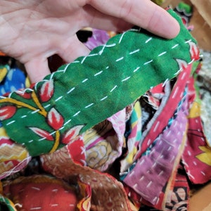 May include: A close-up of a pile of colorful fabric scraps, some of which are sewn together with white thread. The fabric scraps are in a variety of patterns and colors, including green, red, yellow, blue, and pink.