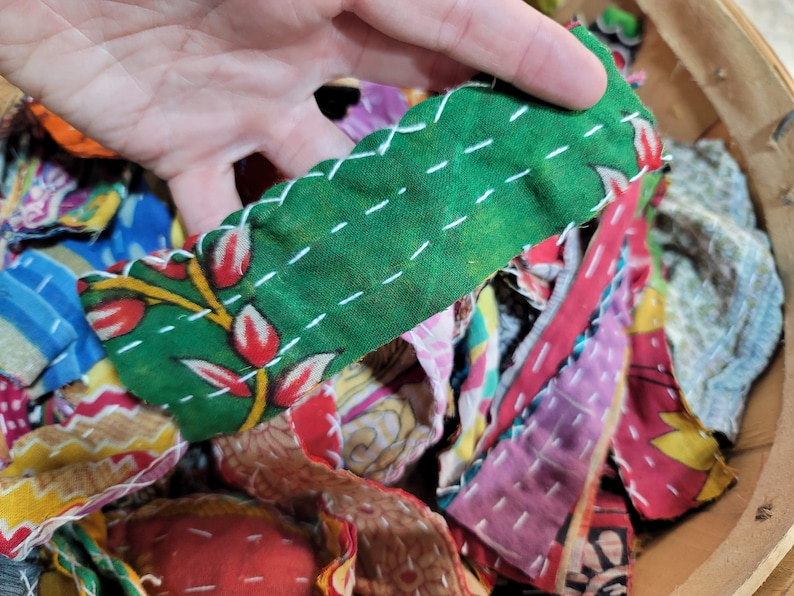May include: A close-up of a pile of colorful fabric scraps, some of which are sewn together with white thread. The fabric scraps are in a variety of patterns and colors, including green, red, yellow, blue, and pink.