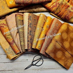 May include: An assortment of patterned fabric pieces in gold, orange, and beige hues. The fabrics display floral and geometric designs. A pair of scissors lies on the wooden surface. The text "Amour Fabriques" is visible on some of the fabrics.