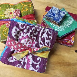 May include: A stack of colorful fabric squares with intricate patterns and stitching. The fabric is tied together with a ribbon and a knot. The fabric squares are arranged in a pile, with the top square being a purple and white floral design.