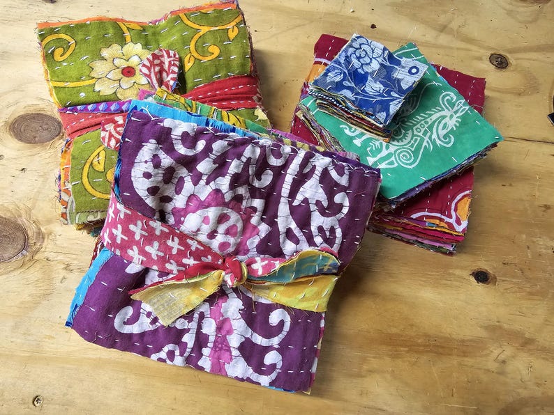 May include: A stack of colorful fabric squares with intricate patterns and stitching. The fabric is tied together with a ribbon and a knot. The fabric squares are arranged in a pile, with the top square being a purple and white floral design.