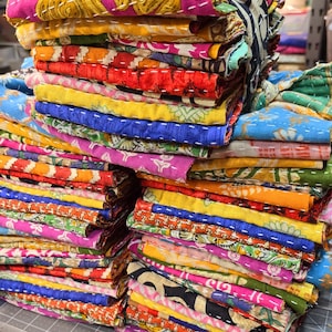 May include: A stack of colorful fabric squares with intricate patterns and stitching. The fabric is folded neatly and arranged in a pile. The fabric is a mix of different colors, including pink, blue, yellow, green, and red.