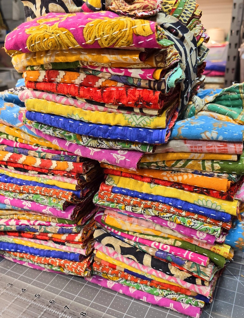 May include: A stack of colorful fabric squares with intricate patterns and stitching. The fabric is folded neatly and arranged in a pile. The fabric is a mix of different colors, including pink, blue, yellow, green, and red.