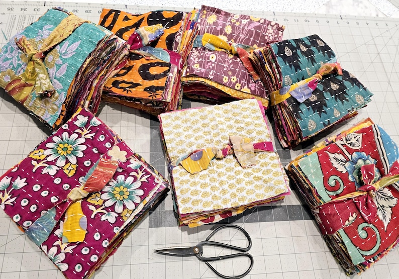 May include: Stacks of colorful fabric squares, tied with twine, are arranged on a gray and white checkered surface. The fabric squares are made of kantha, a type of Indian hand-stitched quilting. A pair of black scissors is lying on the surface.