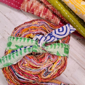 May include: A roll of colorful fabric scraps tied with a green and blue patterned fabric ribbon. The fabric scraps are layered and rolled into a tight bundle.