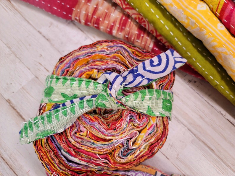 May include: A roll of colorful fabric scraps tied with a green and blue patterned fabric ribbon. The fabric scraps are layered and rolled into a tight bundle.