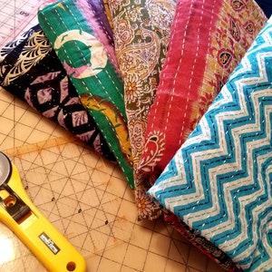 May include: A stack of six colorful fabric squares with different patterns, including paisley, zigzags, and floral designs. The fabric squares are arranged in a fan shape and are likely intended for quilting or crafting.