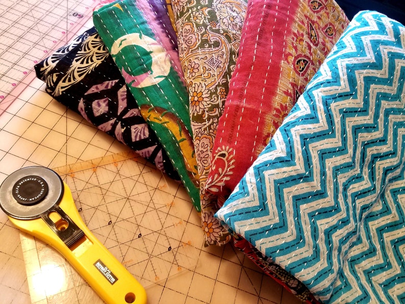 May include: A stack of six colorful fabric squares with different patterns, including paisley, zigzags, and floral designs. The fabric squares are arranged in a fan shape and are likely intended for quilting or crafting.