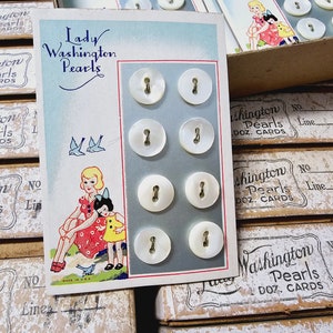 May include: A card featuring the text "Lady Washington Pearls" and a display of six white pearl buttons with two holes each. The card also includes a cartoon illustration of two girls in dresses and birds.