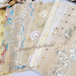 May include: A collection of sheer fabric samples in various shades of cream and beige, embellished with intricate embroidery, sequins, and floral patterns. Some fabrics feature zigzag and floral borders. The text "Amour Fabriques" is visible.