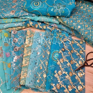 May include: A selection of turquoise fabrics with gold and silver detailing. The textiles display elaborate patterns of sequins and embroidery. The image includes scissors and the text "AmourFabriques".