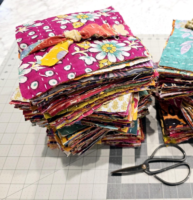 May include: A stack of colorful fabric squares, likely for quilting or patchwork, with a pair of scissors on a cutting mat.