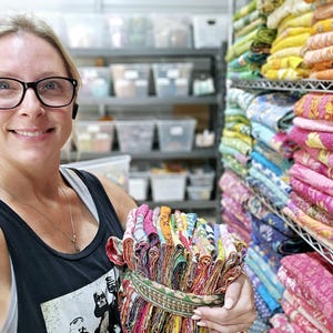 May include: A woman holds a stack of colorful fabric squares, likely for quilting or crafting. The fabric is folded and tied together with a brown ribbon. The background shows shelves filled with similar fabric stacks.