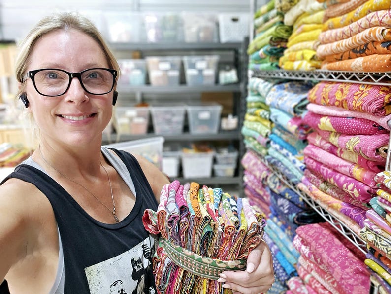 May include: A woman holds a stack of colorful fabric squares, likely for quilting or crafting. The fabric is folded and tied together with a brown ribbon. The background shows shelves filled with similar fabric stacks.