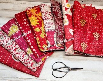 May include: A stack of red, yellow, and white kantha quilts with intricate embroidery and stitching. The quilts are folded and arranged on a white wooden surface. The text "Amour Fabriques" is visible on the quilts.