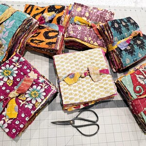 May include: A collection of colorful fabric squares, each tied with a ribbon, are arranged on a gray grid surface. The squares are made of different patterns and colors, including floral, geometric, and paisley designs. A pair of black scissors is also visible in the image.
