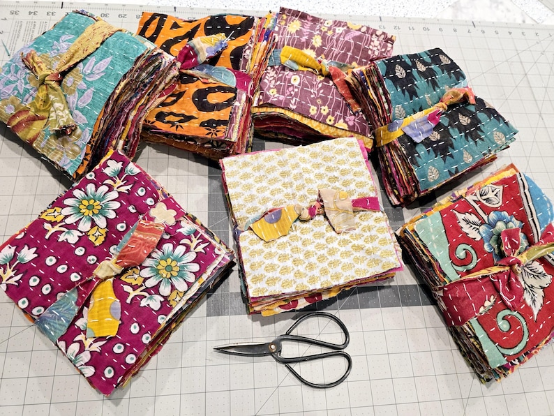 May include: A collection of colorful fabric squares, each tied with a ribbon, are arranged on a gray grid surface. The squares are made of different patterns and colors, including floral, geometric, and paisley designs. A pair of black scissors is also visible in the image.