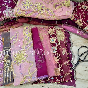 May include: Assortment of pink and burgundy fabric scraps with gold and silver embroidery and sequins. The fabric scraps are arranged in a row on a wooden surface. A pair of black fabric scissors is lying on the surface.