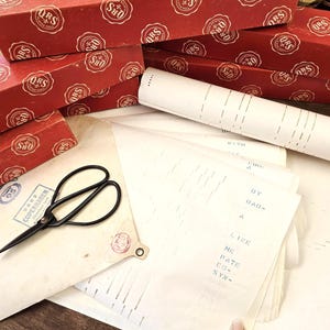 Op de afbeelding: Een verzameling vintage items, waaronder rode dozen met witte cirkelvormige logo's, bladmuziek, een opgerold papier en een zwarte schaar. De bladmuziek heeft tekst en stippellijnen. De dozen zijn opgestapeld en de schaar is open.