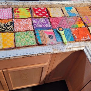 May include: A collection of colorful fabric squares arranged on a countertop, ready to be sewn together to create a quilt. The squares are made of various fabrics, including silk, cotton, and linen, and feature a variety of patterns and designs. A ruler and rotary cutter are also visible on the countertop.
