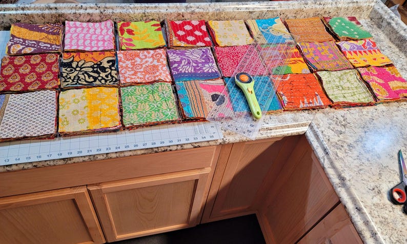 May include: A collection of colorful fabric squares arranged on a countertop, ready to be sewn together to create a quilt. The squares are made of various fabrics, including silk, cotton, and linen, and feature a variety of patterns and designs. A ruler and rotary cutter are also visible on the countertop.