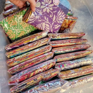 May include: A stack of colorful fabric squares wrapped in plastic, some with floral patterns, others with geometric designs. The fabric squares are arranged in a clear plastic container.